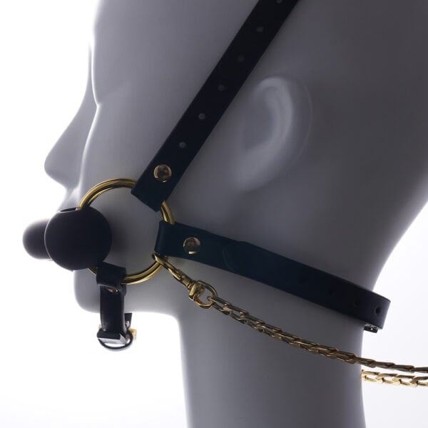 Adjustable headrest harness and gag in black leather, adorned with gold rings and chains, is presented on a white mannequin standing still in profile, with a light background.