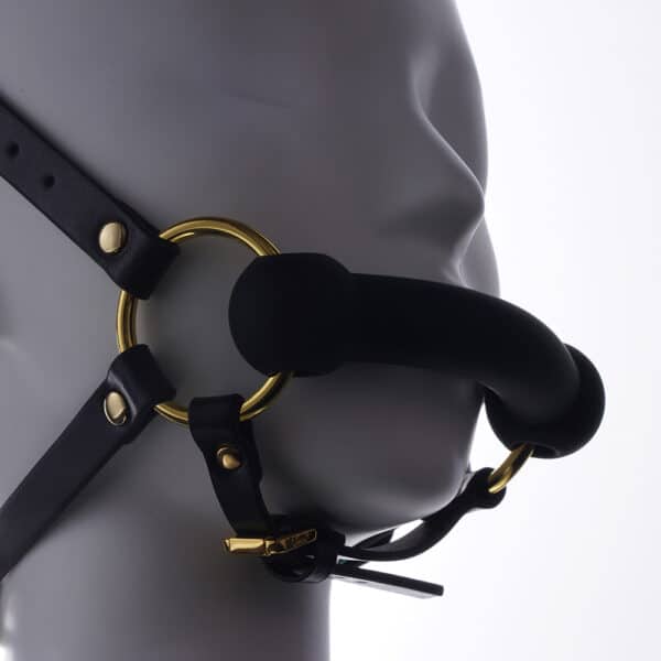 Adjustable headrest harness and gag in black leather, adorned with gold rings and chains, is presented on a white mannequin standing still in profile, with a light background.