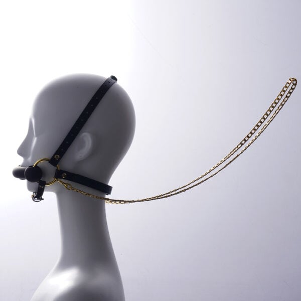 Adjustable headrest harness and gag in black leather, adorned with gold rings and chains, is presented on a white mannequin standing still in profile, with a light background.