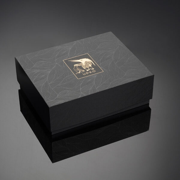 Le packaging UPKO, noir et doré, est disposé en diagonale sur un fond noir brillant qui capte et reflète la lumière blanche.