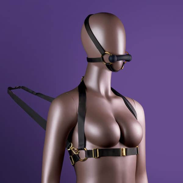 Adjustable head and body harness, with black leather gag and reins attached to the chest, adorned with gold rings and chains, is presented on an immobile mannequin.