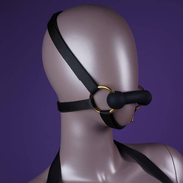 Adjustable head and body harness, with black leather gag and reins attached to the chest, adorned with gold rings and chains, is presented on an immobile mannequin.