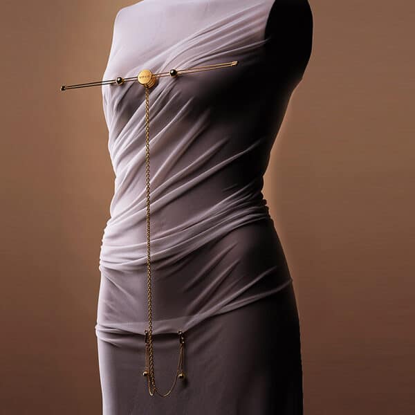 Foto vor neutralem Hintergrund von einem Frauenkörper, der ein weißes, drapiertes Outfit trägt. Sie trägt ein Accessoire mit Nippelklemme und goldener Klitoriskette.