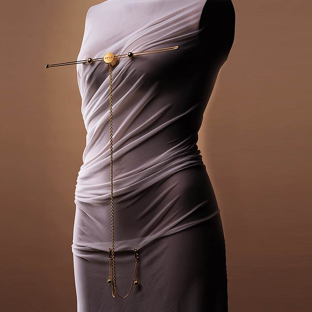Photo sur fond neutre d'un corps de femme portant une tenue blanche et drapée. Elle porte un accessoire barre pince à tétons et chaine clitoridienne dorée.