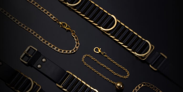 Black background photograph of a black bondage set, showing a belt and gold chains.