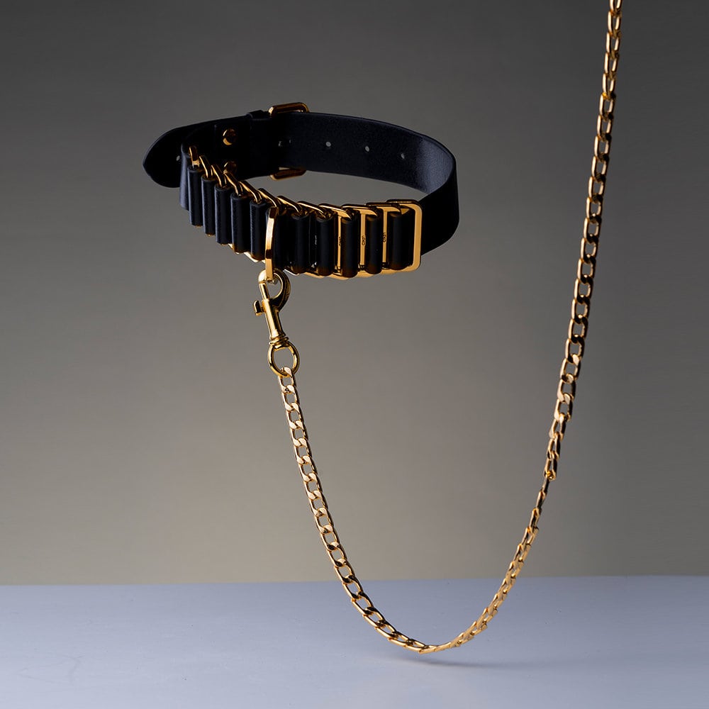 Photographie sur fond gris montrant un collier noir avec des détails dorés, relié à une chaine dorée.