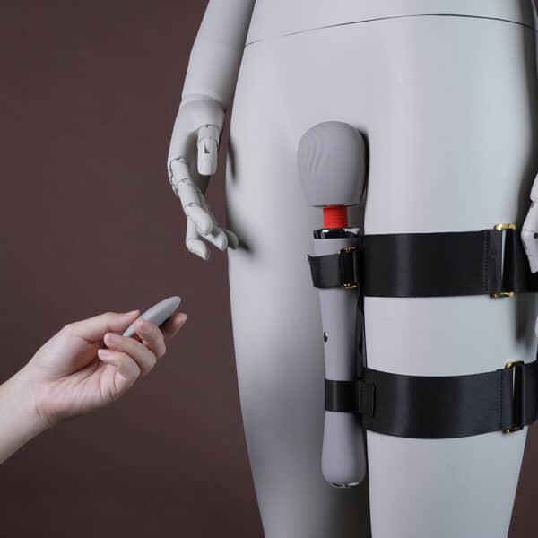 Image showing a leg to which a set with a black harness and a white vibrator is attached. One hand holds the remote control.
