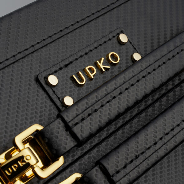 Photographie en zoom de la poignée de la malle noire en cuir. Il y a deux poignées avec des détails dorés. Au dessus le nom de la marque est inscrit.
