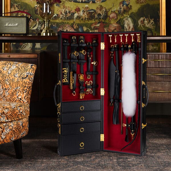 Photograph in a room with animal decor, a black trunk with red interior is in the center. It contains various leather accessories with gold details.
