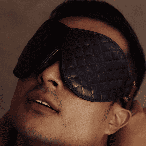 Photograph of a man's face. He is blindfolded with a black leather fleece blindfold.