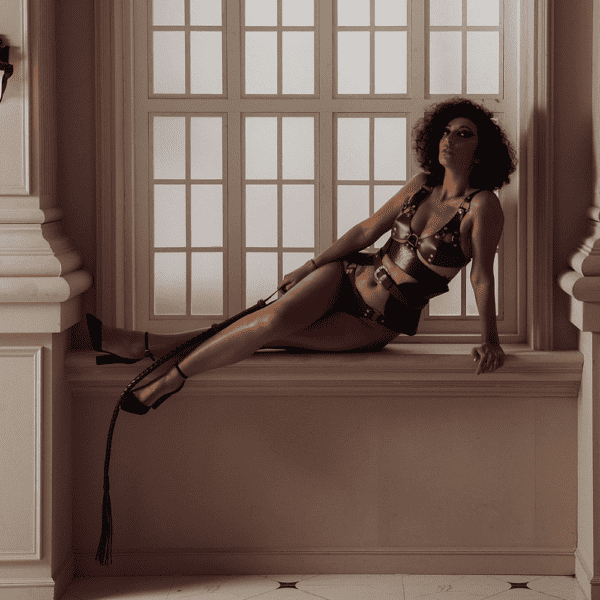 Photograph of a woman lying on a white windowsill. She is wearing a leather lingerie set. A bralette, belt and panties. She also has a leather whip in her hand.