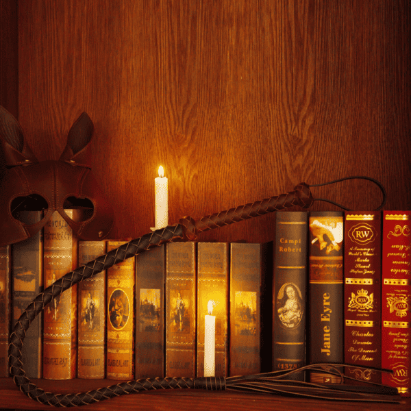 Photographie de près d'un fouet tressé en cuir et d'un masque en cuir marron. LE tout disposé sur des livres et accompagné de bougie blanches.