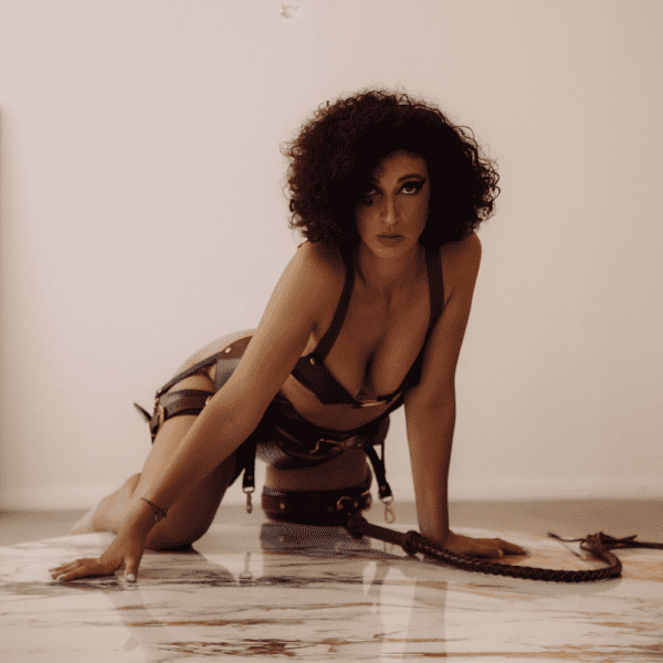 Photograph of a woman on all fours on the floor. She has a brown leather whip in front of her and is wearing a brown lingerie set.