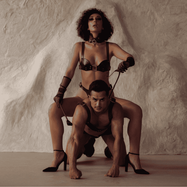 Photogaphie d'une femme et d'un homme portant de la lingerie en cuir marron. La femme est assise sur le dos de l'homme qui lui est à quatre pates. La femme porte un soutien gorge en cuir marron, elle a des gants en mesh noire et dans sa main une cravache. L'homme lui porte un harnais en cuir marron.