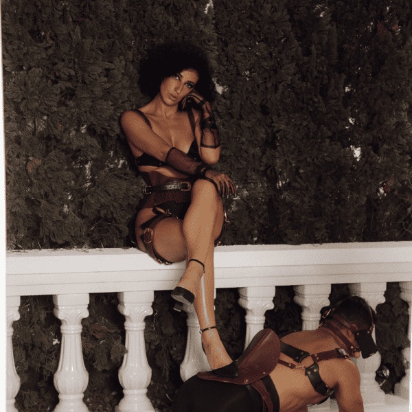 Dark photograph of a dominant woman in equestrian leather lingerie sitting on a low wall, her submissive on all fours on the ground and her feet resting on the saddle on her back.