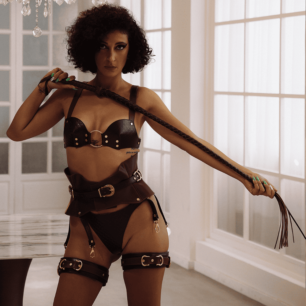 Photograph of a woman wearing a brown lingerie ensemble and holding a whip in front of her. 