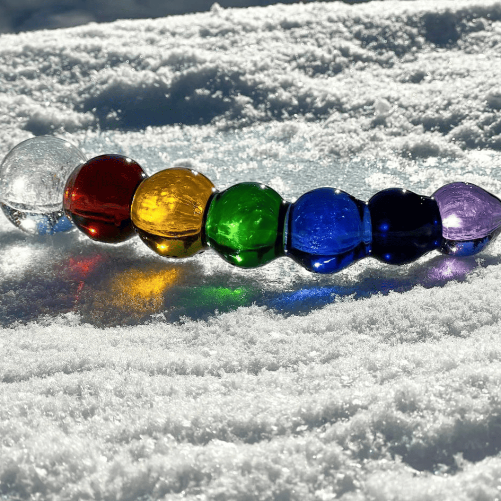 Fotografía de un consolador en la nieve sobre fondo blanco, representando un arco iris, coloreado de blanco, rojo, amarillo, verde, azul, azul marino y violeta, un festival de confeti y colores.