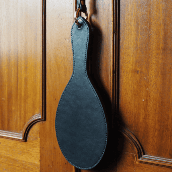 Photograph of the Paddle Fessée Cuir hanging from a wooden cabinet