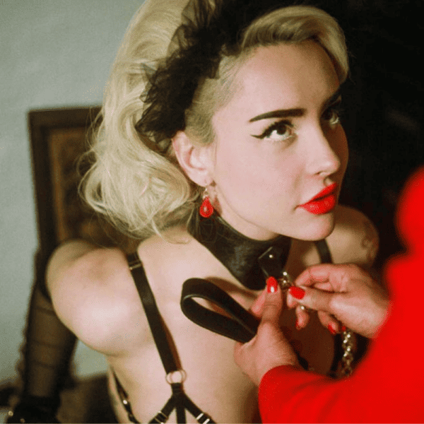 Blonde woman with bright lipstick and piercing eyes, is leashed with black leather chocker.