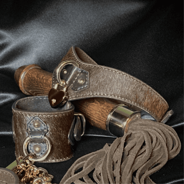 Brown leather whip with wooden handle in the shape of a dildo with leash collar and leather handcuffs set against a black textile background.