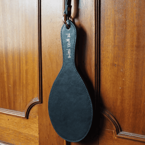 Photograph of the Paddle Fessée Cuir hanging from a wooden cabinet