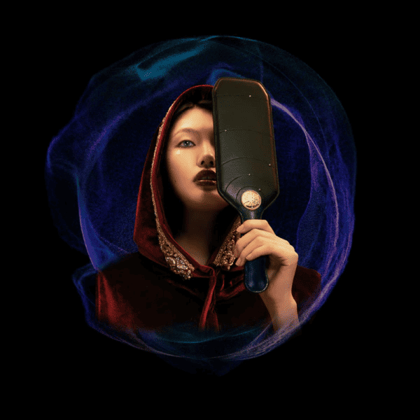 Black background photograph of a female witch holding a black baked paddle in her hand, wearing a red cape and gold embroidery.