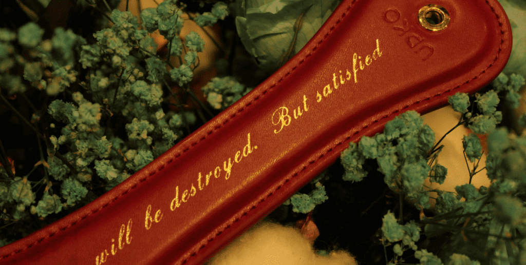 Photograph of the Upko Rouge leather paddle with gold lettering that reads "You will be destroyed. but satisfied". The red paddle is set in a bouquet of blue flowers.