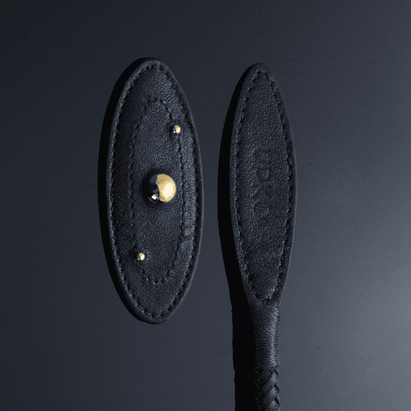 Photographie zoomé sur le bout du fouet posé sur un fond noir réfléchissant la lumière , détails de boules en or, coutures parfaites, nom de la marque gravé dans le cuir noir et manche tressé