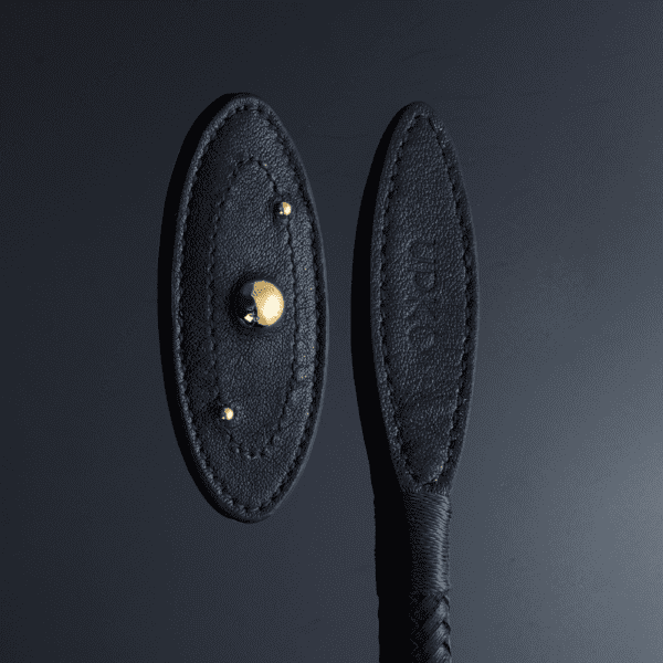 Zoom in on the tip of the whip set against a light-reflecting black background, gold ball details, perfect stitching, brand name engraved in the black leather and braided handle.