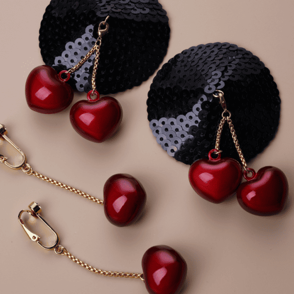 Packshot on beige background of the cherry heart-shaped clitoral jewelry with red heart and gold chain and hook and black glitter base with clitoral jewelry with bells