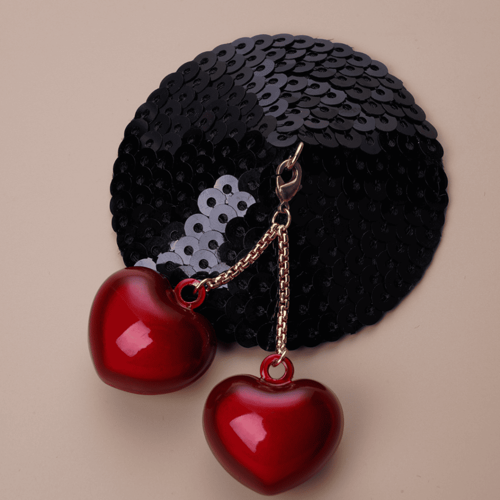 Packshot photograph on a beige background of the cherry heart-shaped cache-tétons with gold chain and hook and black glitter base.