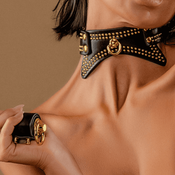 Young woman posing against a brown background. She is wearing a black leather necklace with gold studded details.