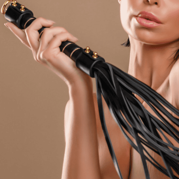 Young woman posing on a brown background. She is holding a leather whip with leather bangs. The whip has gold metallic details.