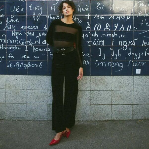 Model wearing Undress Code Go For It bodysuit, posing in front of a wall with writing. With black pants and red shoes.