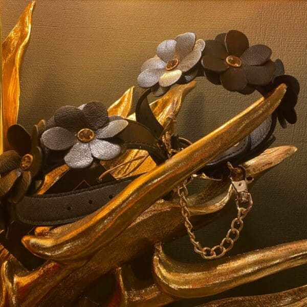 Schwarze Lederhandschellen im Blumendesign, die auf einem goldenen, leuchtenden Dekor liegen.