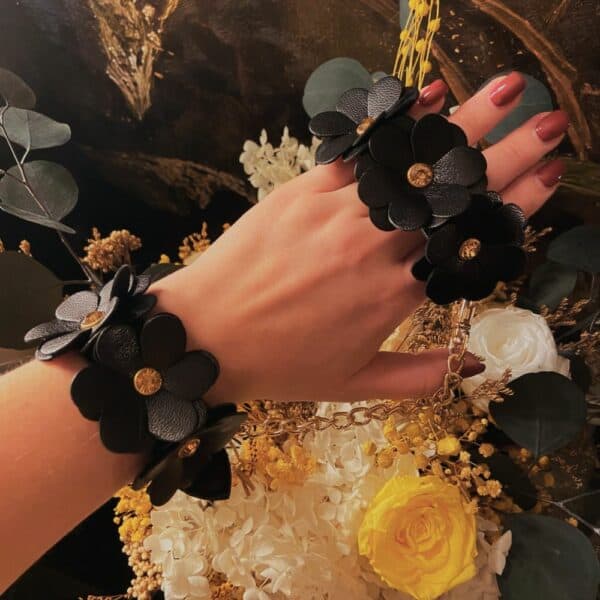 Hand holding the two shiny black leather handcuffs, flower design with gold center, the handcuffs are linked by a gold chain.