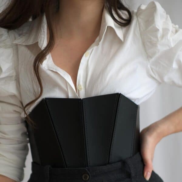 Corset 71458 noir porté par une femme avec une chemise blanche et un pantalon noir. Le corset en cuir pleine fleur rigide accentue la taille et la poitrine avec un décolleté profond, tandis que le système de laçage discret à l'arrière apporte une finition élégante.