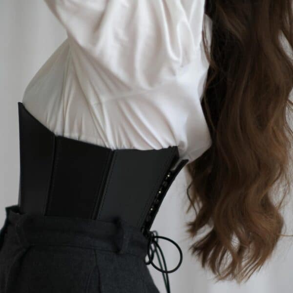 Corset 71458 noir porté par une femme avec une chemise blanche et un pantalon noir. Le corset en cuir pleine fleur rigide accentue la taille et la poitrine avec un décolleté profond, tandis que le système de laçage discret à l'arrière apporte une finition élégante.