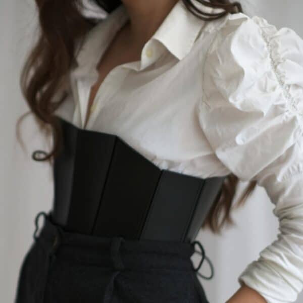 Corset 71458 noir porté par une femme avec une chemise blanche et un pantalon noir. Le corset en cuir pleine fleur rigide accentue la taille et la poitrine avec un décolleté profond, tandis que le système de laçage discret à l'arrière apporte une finition élégante.