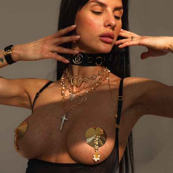 Золотые украшения и накладки на соски QRUCIFIX - BRIGADE MONDAINE - Роскошное белье Paris На женщине несколько золотых ожерелий QRUCIFIX и металлические накладки на соски в форме дисков с подвесками. Она позирует с поднятыми руками и закрытыми глазами. Виден черный чокер с логотипом и черные бретельки нижнего белья.